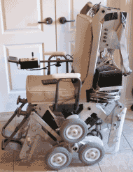 Level Position” shows the side view of the StairMaster with support by the forward minor wheels and rear caster wheels. The rear minor wheels are raised slightly off the floor. The rear caster wheels are hidden. The rear caster skids are shown behind the spider wheel in an almost vertical position.  The front skids are folded up. Leg rest, arm rest with controller unit are shown. The wheelchair has a high seat back with a computer shown behind it.  The batteries are visible below the computer. 